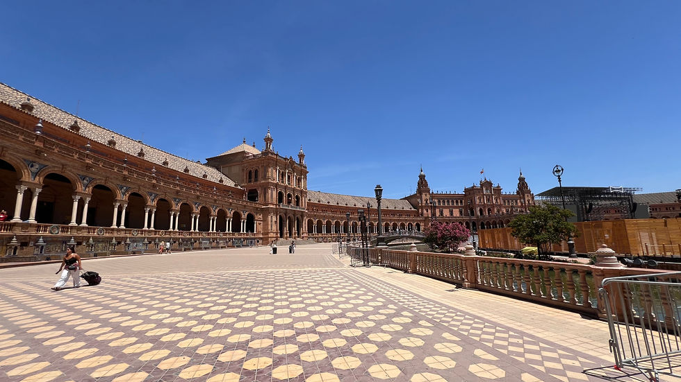 Sunlit ornate Spanish plaza with arcaded buildings, blue sky, and a woman pulling a suitcase across patterned tiles.