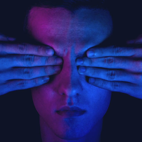 Person covers eyes with hands in a dark setting, lit by purple and blue light. The mood is mysterious and introspective. No text.
