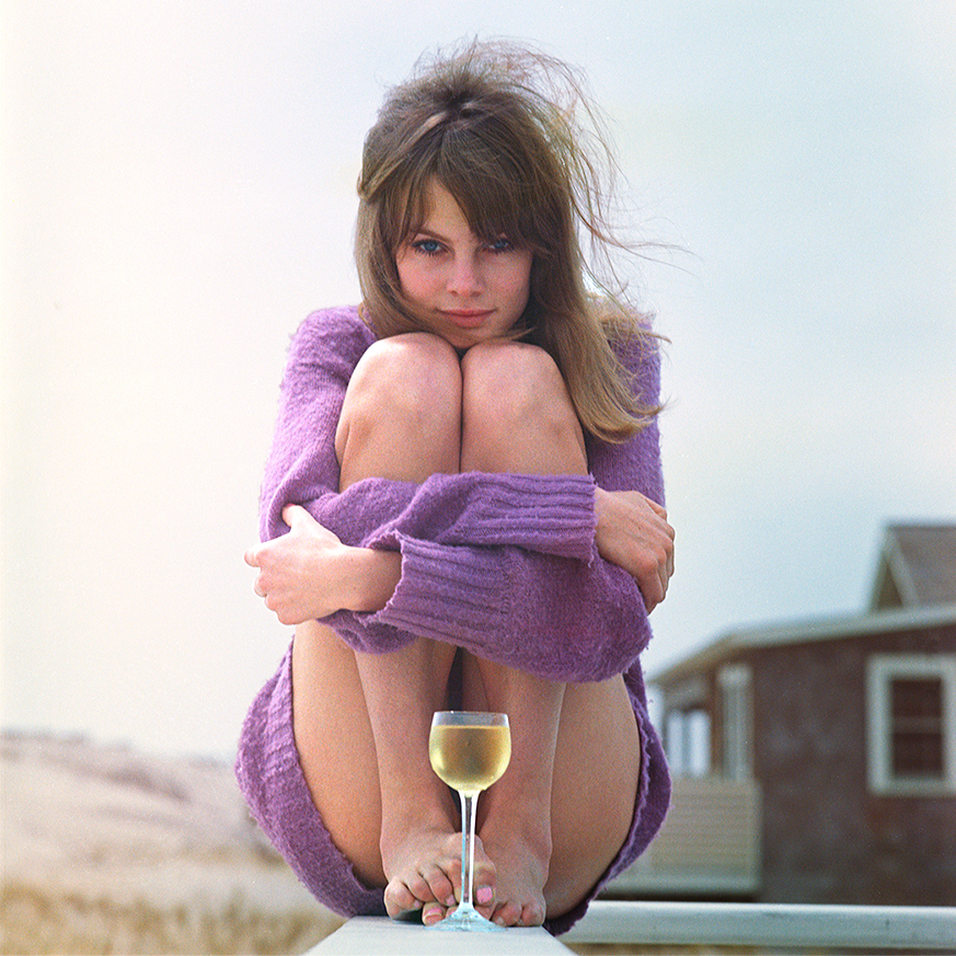 Woman in a purple sweater sits with legs crossed on a rooftop, holding a glass of white wine. Overcast sky and building in background. Mood is serene.