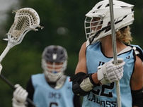 Youth lacrosse players in helmets and numbered jerseys huddle on a field, sticks raised, with trees blurred behind.