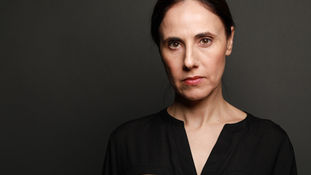 Serious woman in black shirt with zippers against dark background. Her expression is neutral and focused.