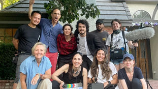 A group of ten people pose cheerfully with film equipment and a clapperboard under a leafy tree, in front of a house with arched windows.