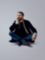 Man with beard sitting cross-legged on floor against a light gradient background, resting chin on hand.