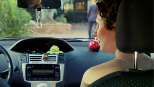 Two people in formal attire sit in a car, looking surprised. The woman appears concerned. Bold text reads "TRAVEL PLANS" at the top.
