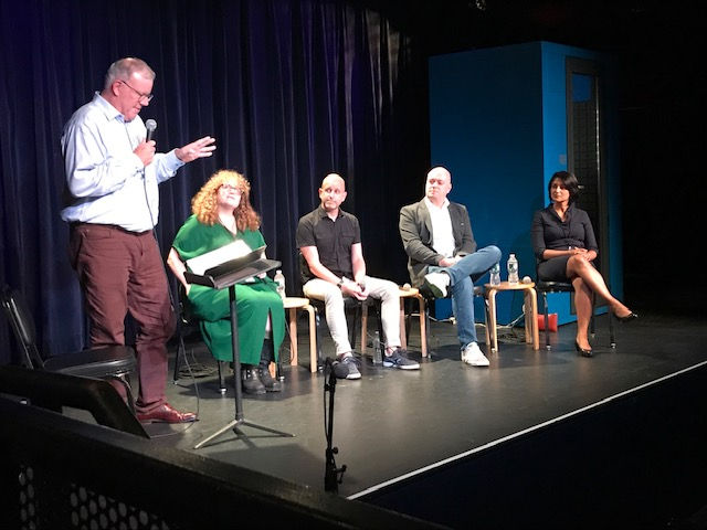 Five people are seated on stage with a blue curtain backdrop. One stands speaking into a microphone. A casual, conversational setting.