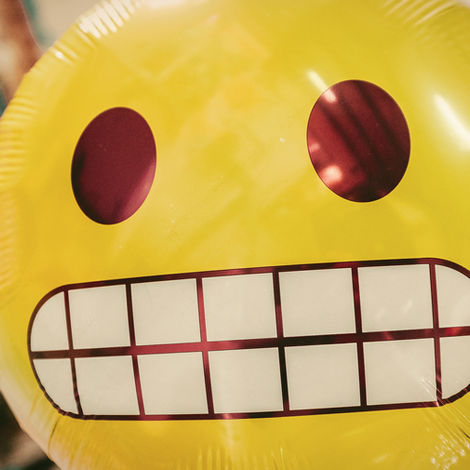 Yellow emoji balloon with large eyes and grid smile, floating indoors. Background has blurred colors, creating a lively atmosphere.