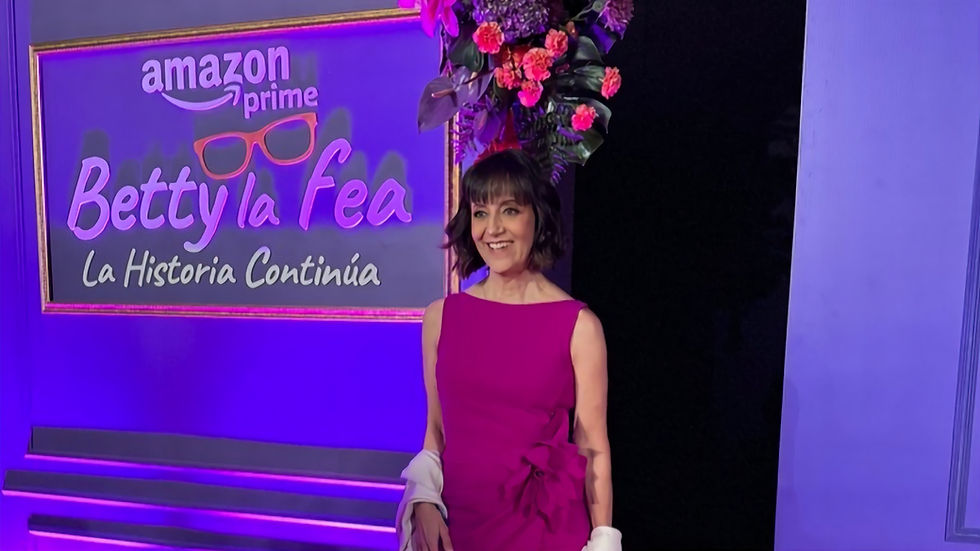 Mujer con vestido fucsia sonríe junto a un letrero de Amazon Prime Betty la fea: La Historia Continúa, bajo luces moradas.