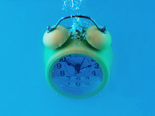 Green alarm clock submerged in clear blue water, creating bubbles. Clock face shows numbers and hands. Bright, dynamic scene.