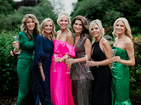 Six women in elegant dresses hold champagne flutes, smiling in a lush garden setting. Dresses are green, pink, blue, and black.