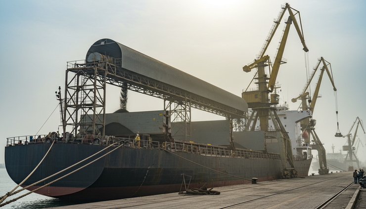 Eye-level view of a bulk cargo vessel being loaded with dark petroleum coke at a port conveyor