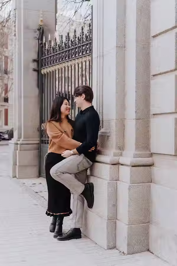 Photo shoot of a couple with the man leaning against a stone pillar and the woman embracing him next to a historic gate in Madrid.