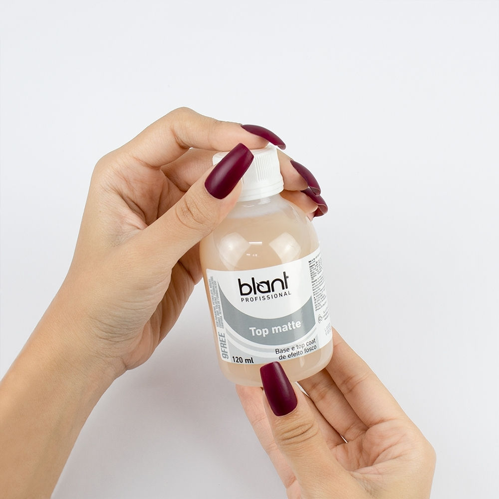 Mãos segurando frasco Blant Top coat, com unhas pintadas de vermelho escuro. Descolados da Web