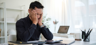 Homem sentado à mesa de trabalho, com notebook aberto, cobrindo o rosto com as mãos em sinal de cansaço e estresse, representando sobrecarga emocional e pressão no ambiente corporativo.