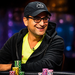Antonio Esfandiari smiling at a poker table with stacks of colorful chips, wearing black glasses, a black cap and dark hoodie.
