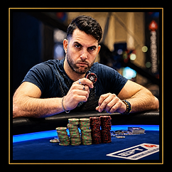 Professional poker player seated at a blue felt table holding a chip, with stacks of tournament chips in front of him.