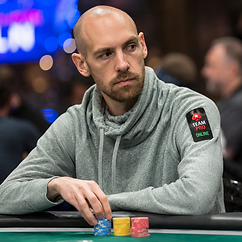 Professional poker player seated at a tournament table in a gray hoodie, stacking blue, yellow, and red poker chips.