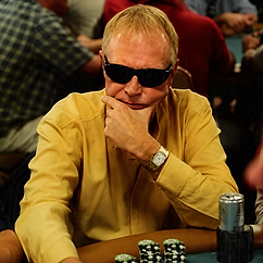 Roger Moore wearing sunglasses and a yellow shirt while seated at a poker table with stacks of chips during a live tournament.