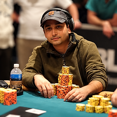 Allen Bari seated at a poker tournament table wearing headphones and a dark cap, with stacks of orange and yellow poker chips in front of him.