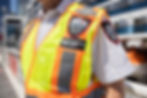 Security guard in a bright yellow vest with orange stripes, labeled "SECURITY," outdoors on a sunny day. Urban background.