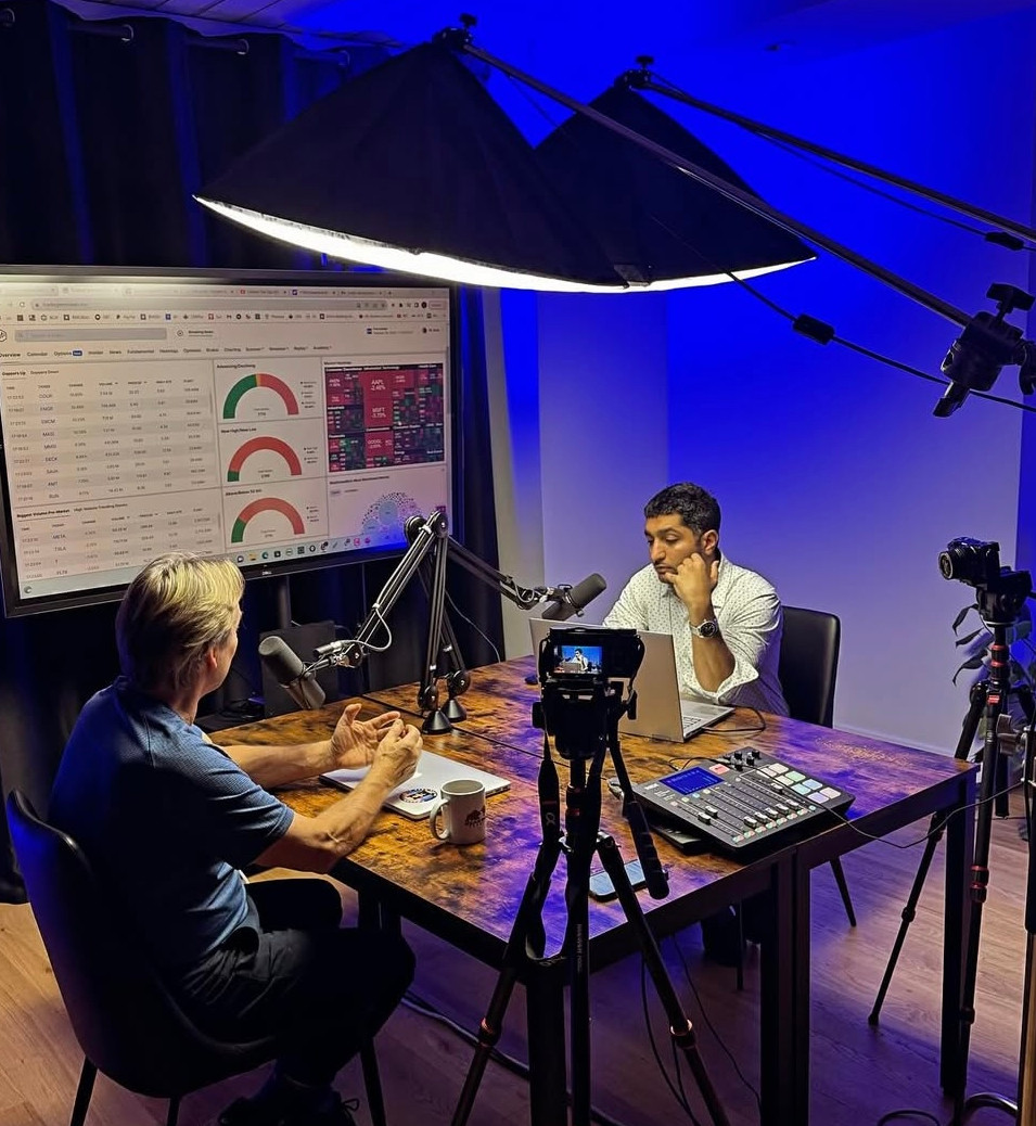 Two men recording a podcast in a studio with trading dashboards on screen and professional lighting setup.