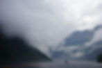 Boat on misty fjord, surrounded by mountains and dense clouds. Gray and green hues dominate, creating a serene, mysterious atmosphere.
