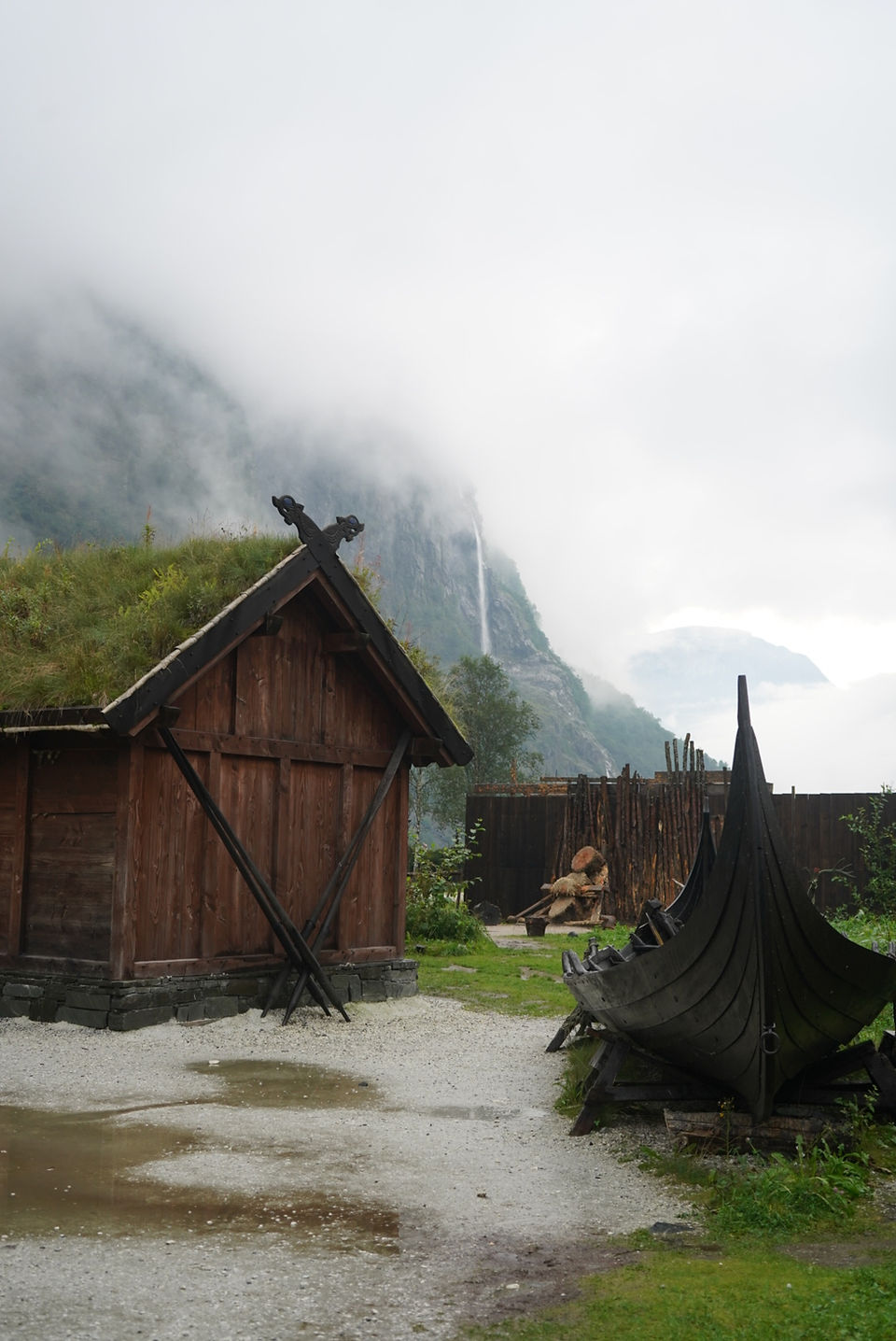 A wooden building with a mossy, grassy roof and a wooden boat in front of clouded mountains and a waterfall.