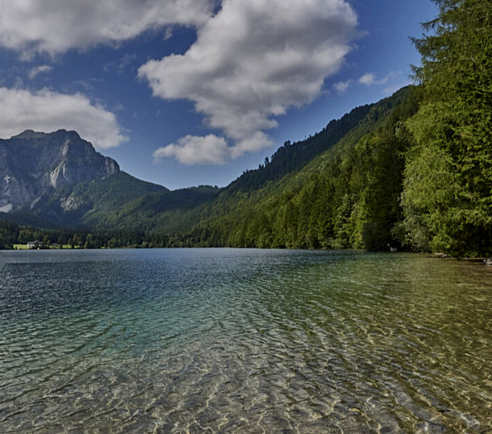 Clear lake with rippling water in the foreground, surrounded by lush green forest and mountains under a cloudy blue sky.