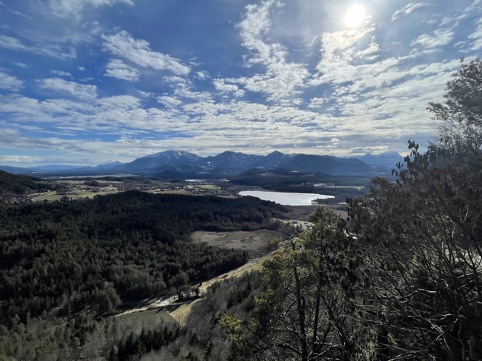 Gozdnat panoramski pogled na gorsko pokrajino, jezero, temen gozd ter sonce na oblaku. Oblačno nebo, miren in slikovit ambient.