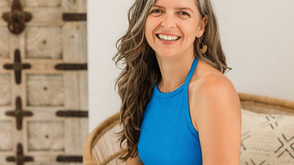 Smiling woman in a vibrant blue top sits comfortably in a woven chair, exuding warmth and joy against a rustic wooden door backdrop.