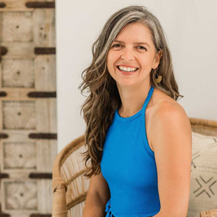 Smiling woman in a vibrant blue top sits comfortably in a woven chair, exuding warmth and joy against a rustic wooden door backdrop.