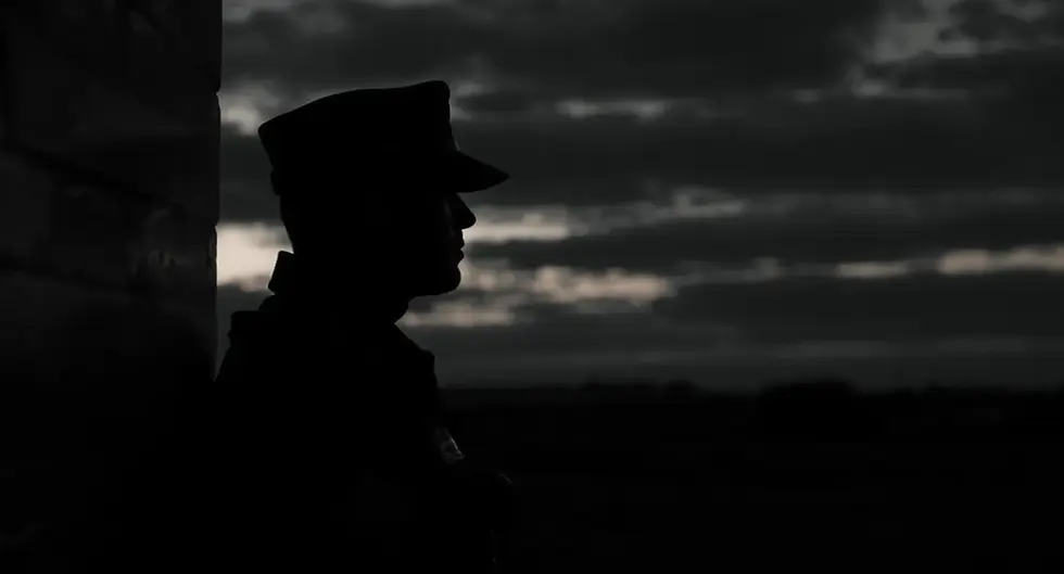 Silhouette of a person in uniform leaning against a wall at sunset. Dark clouds fill the sky, creating a somber, reflective mood.