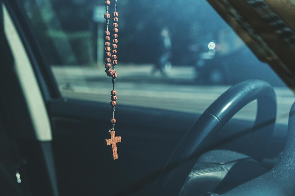 Rosary with wooden cross hanging from rearview mirror in car. Blurred street view outside, with muted colors and a calm mood.