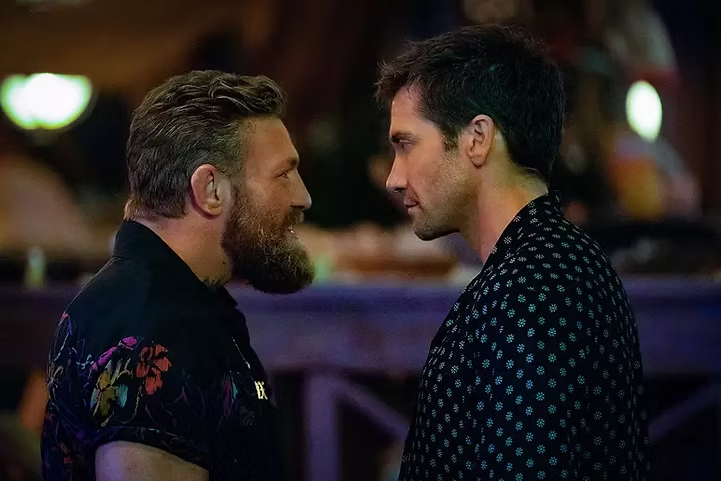 Conor McGregor encara Jake Gyllenhaal em uma cena intensa do filme "Matador de Aluguel" (2024). Os dois personagens estão frente a frente em clima de tensão, em um ambiente noturno e vibrante, típico de um bar ou clube.