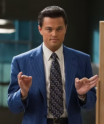 Leonardo DiCaprio caracterizado como Jordan Belfort no filme O Lobo de Wall Street, usando terno azul e fazendo gesto icônico com as mãos em cena de escritório.