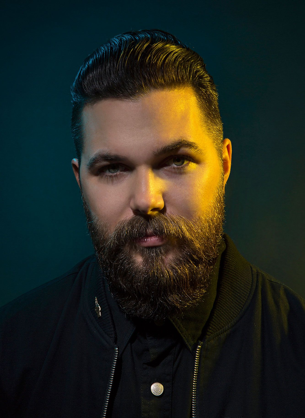 Retrato de Robert Eggers de barba cheia e cabelo penteado para trás, usando jaqueta preta em um fundo escuro com iluminação dramática em tons de verde e amarelo.