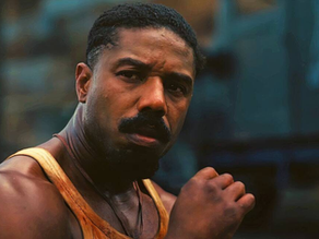 Michael B. Jordan com expressão séria e suado, vestindo regata manchada em cena tensa do filme “Pecadores”, com fundo industrial desfocado.