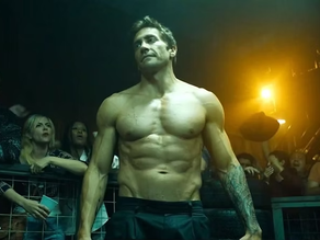 Jake Gyllenhaal sem camisa exibe físico definido em cena de luta clandestina no filme "Matador de Aluguel" (2024), cercado por uma multidão animada e luzes dramáticas ao fundo.