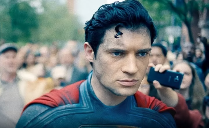 Superman 2025 com expressão séria em meio a multidão filmando com celular – traje azul e vermelho, cacho clássico na testa, atmosfera urbana