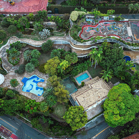 Vista aérea de um parque urbano com piscina azul, plantas tropicais e caminhos sinuosos. Telhados vermelhos e rua cinza ao redor. Ambiente tropical.