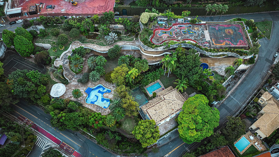 Vista aérea de um parque urbano com piscina azul, plantas tropicais e caminhos sinuosos. Telhados vermelhos e rua cinza ao redor. Ambiente tropical.