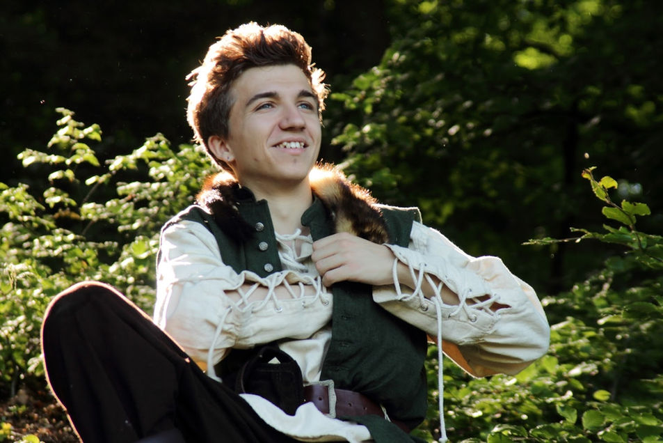 Ein junger lächelnder Mann sitzt im Wald. Er trägt ein Fantasy-Kaufmannskostüm mit Pelzkragen und greift in seine Innentasche. Die Sonne beleuchtet ihn von hinten.