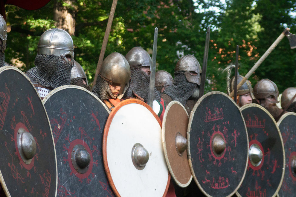 Eine Reihe von 9 Wikingerkriegern in voller Rüstung mit Waffen wie Schwertern und Äxten halten ihre Rundschilde hoch, während sie auf den Angriff warten.