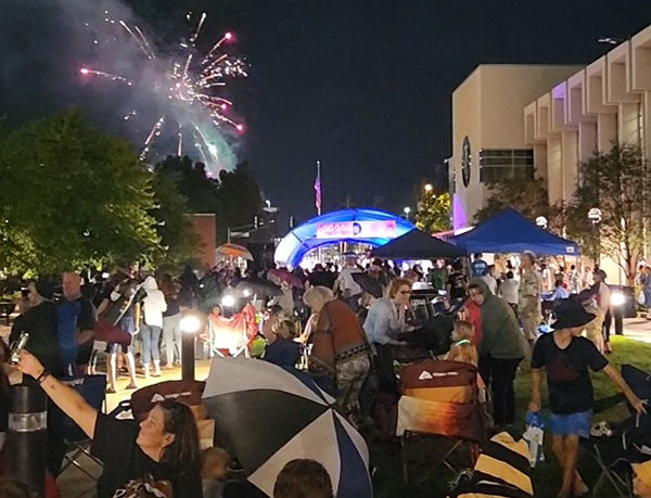 People sitting outside in the evening listening to a rock concert. Fireworks from the Tin Caps are going off in the night sky in the background. 