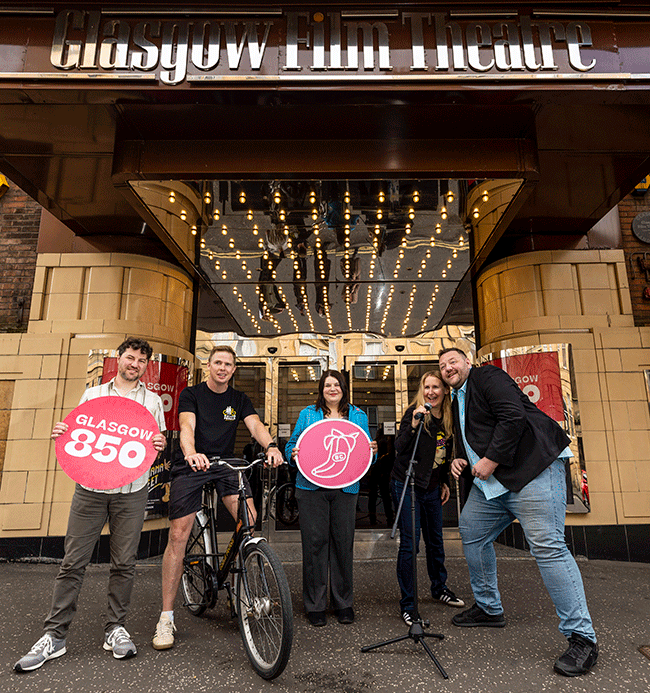 Five people outside Glasgow Film Theatre hold signs and a microphone; one has a bike. The background shows bright marquee lights.
