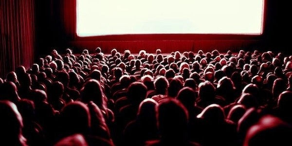 Audience in a theater facing a lit screen. The scene is cast in red tones, creating a dramatic and focused atmosphere.