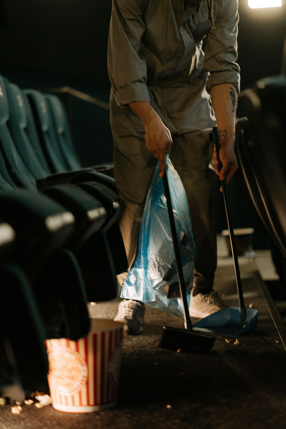 Person cleaning up a movie theater aisle, sweeping popcorn into a dustpan. An empty striped popcorn bucket is on the floor. Dim lighting.