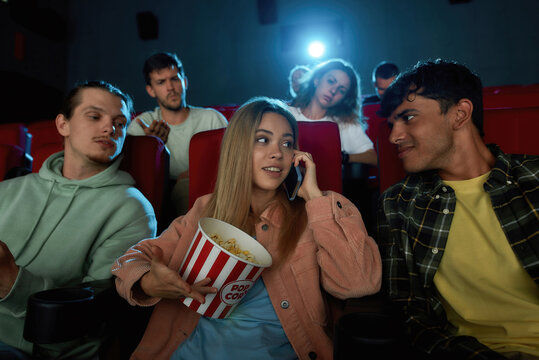 Woman with popcorn talks on phone in a movie theater, surrounded by others looking annoyed. Dim lighting, red seats, casual attire.