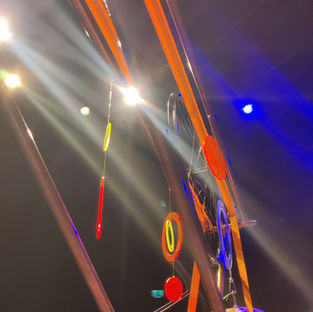 A close up of a structure made of long copper pipes and orange cargo straps, with colourful decorative discs hanging from it. We are looking up at it and stage lights are visible above it, creating white and blue flares of light against a black background.