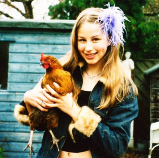 A white teenage girl with long light brown hair, wearing a denim jacket with furry trim, a black top and a purple feathery hair accessory. She is standing in front of trees and a blue garden shed, smiling and holding a brown chicken. The chicken is also looking at the camera.