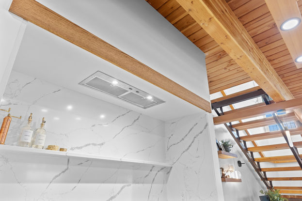 A spacious, modern industrial-style open-concept living area featuring polished concrete floors and warm wood-beamed ceilings with exposed ductwork. The space includes a contemporary kitchen with white cabinetry and a curved breakfast bar, a dining area with a built-in banquette, and a staircase leading to an upper level